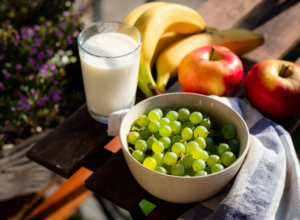Ciotola di uva verde con bicchiere di latte, mele e banane su tavolo all’aperto. Nutrizione